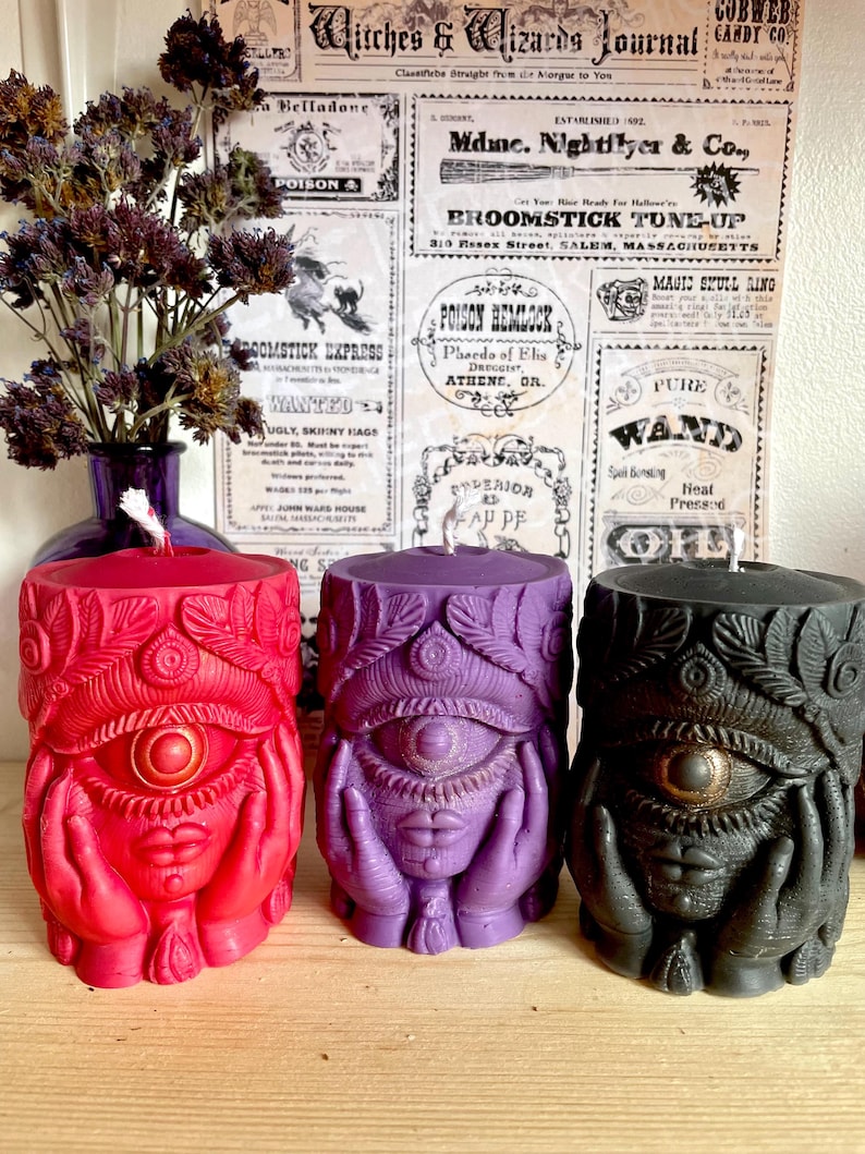 May include: Three pillar candles in red, purple, and black, each with a detailed eye design and hands. The candles have a textured surface and are part of a witchy or mystical theme.