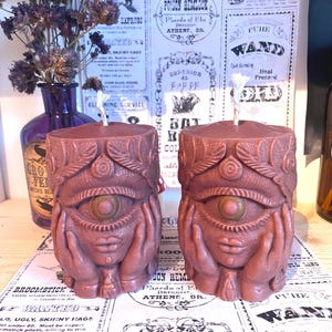 May include: Two brown candles with an eye and hands design. The candles feature a detailed carved design with an eye in the centre and hands on each side. The candles are on a table with vintage-style paper in the background.