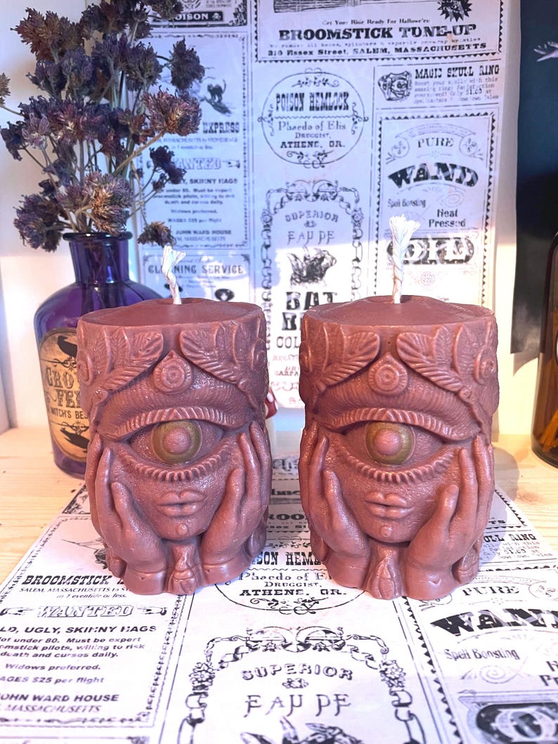 May include: Two brown candles with an eye and hands design. The candles feature a detailed carved design with an eye in the centre and hands on each side. The candles are on a table with vintage-style paper in the background.