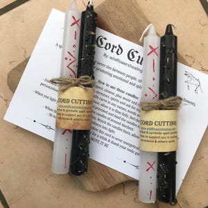 May include: Two black and white candles with red markings and a twine bow. The candles are labelled "Cord Cutting" and have a description of their use. The candles are on a wooden surface.