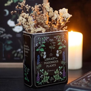 Könnte beinhalten: Eine schwarze, buchförmige Keramikvase mit getrockneten Blumen. Die Vase ist mit botanischen Illustrationen und dem Text "Library of Poisonous Plants" verziert. Ein Totenkopf- und gekreuzte Knochen-Symbol ist ebenfalls vorhanden.