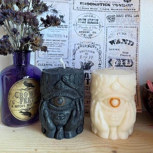 May include: Two pillar candles with eye designs, one black and one cream, sit on a wooden surface. A purple bottle with a label that reads "Crow's Feet, A Witch's Best Friend" is to the left. The background features vintage-style advertisements.
