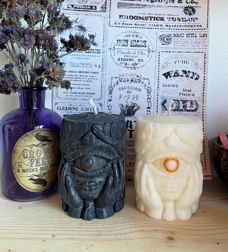 May include: Two pillar candles with eye designs, one black and one cream, sit on a wooden surface. A purple bottle with a label that reads "Crow's Feet, A Witch's Best Friend" is to the left. The background features vintage-style advertisements.
