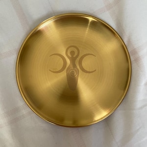 May include: A circular, gold-coloured metal tray with a brushed finish. It has an engraved design of a central figure with crescent moons. This decorative tray is a stylish item.