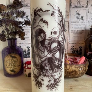 May include: A cream-coloured pillar candle with a black and white illustration of Jack Skellington and Sally. The design includes pumpkins, bats, and a spooky forest scene, ideal for Halloween decorations and themed settings.