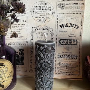 May include: A dark grey pillar candle with a white wick, adorned with a repeating pattern of stars and crescent moons. The candle is set against a vintage newspaper backdrop, with dried flowers in a purple bottle to the left.