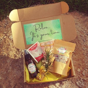 May include: A brown cardboard box with a yellow tissue paper lining. The box contains a variety of self-care items, including a bottle of body spray, a bag of bath salts, a tea bag, and a wooden stick. The box also includes a card with the text "Relax... It's your time..." and a website address.