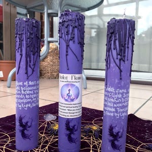 May include: Three purple pillar candles with dripped wax. Each candle has a label with text and a phoenix illustration. The candles are on a purple cloth with dried flowers.