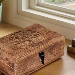 Könnte beinhalten: Eine rechteckige Holzschatulle mit natürlicher brauner Maserung und einem kreisförmigen, geschnitzten Lebensbaum-Motiv auf dem Deckel. Die Schatulle ist mit einem kleinen schwarzen Metallverschluss an der Vorderkante versehen, der sie sicher verschließt.