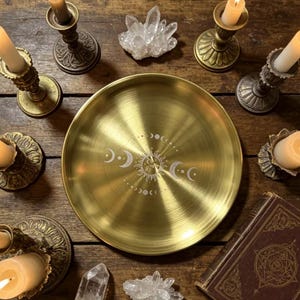 May include: A circular brass tray featuring an etched sun and moon phase design in the centre, surrounded by six antique-style metal candle holders with lit cream-coloured taper candles, two clear quartz crystal clusters, and a vintage book.