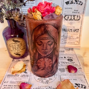 Puede incluir: Una vela cilíndrica marrón con un retrato de mujer, coronada con una rosa rosa, flores secas y cristales. La vela tiene un diseño de estilo vintage. El fondo incluye recortes de periódicos antiguos y un jarrón morado.