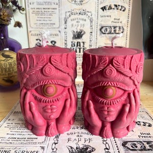 May include: Two pink candles with an eye, hands, and lips design. The candles have a detailed, carved appearance and are placed on a surface with vintage-style paper. The candles have a wick at the top.
