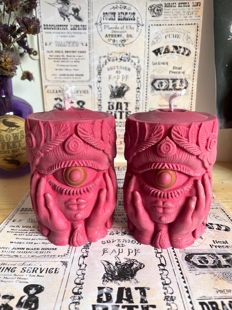 May include: Two pink candles with an eye, hands, and lips design. The candles have a detailed, carved appearance and are placed on a surface with vintage-style paper. The candles have a wick at the top.