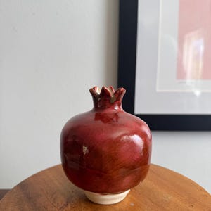 May include: A ceramic pomegranate-shaped vase with a glossy, deep red finish. The vase has a small, textured crown and a white base. The decorative piece is set on a wooden surface, with a framed artwork in the background.