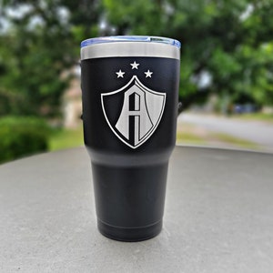 May include: Black stainless steel tumbler with a white engraved shield and the letter "A" inside. There are five stars above the shield.