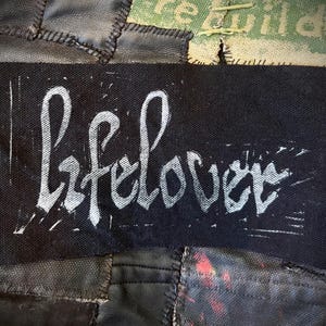 May include: A black fabric patch with the word "lifelover" in white, handwritten-style lettering. The patch is rectangular and appears to be sewn onto a patchwork background of black leather and other fabrics.