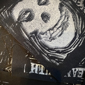 May include: Black and white fabric patches featuring a detailed illustration of a skull-like face with closed eyes and a wide grin. The patches have a textured appearance and are likely for clothing or accessories.