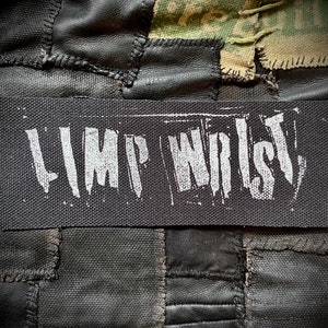 May include: Black rectangular patch with the words "LIMP WRIST" in white, stylized lettering. The patch is set against a background of black fabric and leather patches, with a camouflage fabric section visible.