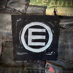 May include: A black square patch with a white, distressed-style letter "E" inside a circle. The patch is sewn onto a patchwork background of various fabrics, including leather and canvas.
