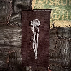 May include: A dark brown fabric patch with a white, stylized illustration of a structure with vertical lines. The patch is sewn onto a patchwork background of brown leather and canvas.
