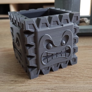 Op de afbeelding: Een grijze 3D-geprinte plantenbak in de vorm van een blok met een cartoon gezicht. Het gezicht heeft twee grote ogen, een brede mond en een gefronste wenkbrauw.