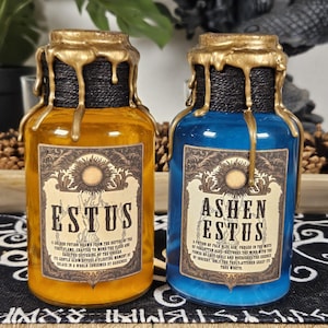 Può includere: Due bottiglie decorative in vetro, una ambrata e una blu, con colature di cera color oro e spago nero attorno al collo. Ogni bottiglia ha un'etichetta con la parola "ESTUS" o "ASHEN ESTUS" e testo decorativo.