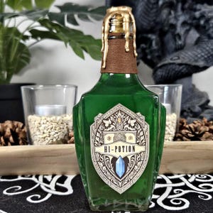 Op de afbeelding: Een groene glazen fles met een gouden waszegel en een label met de tekst "HI-POTION". Het label heeft een sierlijk ontwerp en een blauw kristal. De fles bevat een groene vloeistof. Het ontwerp van de fles doet denken aan een drankfles.