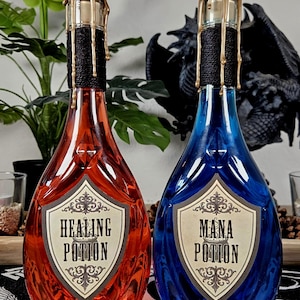 Könnte beinhalten: Zwei dekorative Zaubertrankflaschen, eine rote und eine blaue, mit goldfarbenen Wachssiegeln. Die rote Flasche ist mit "Healing Potion" und die blaue Flasche mit "Mana Potion" beschriftet. Beide Flaschen haben schwarzen Bindfaden um den Hals.