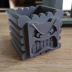 Könnte beinhalten: Ein grauer 3D-gedruckter Pflanztopf in Form eines Blocks mit einem missmutigen Gesicht. Der Topf hat eine strukturierte Oberfläche und ist für kleine Pflanzen oder andere Dekorationsartikel gedacht.