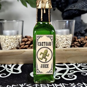 Könnte beinhalten: Eine grüne Glasflasche mit einem schwarzen Etikett mit der Aufschrift "CACTUAR JUICE" und einer Cartoon-Kaktusfigur. Die Flasche ist mit einem schwarzen Verschluss und goldenem Wachs versiegelt. Dekoration mit Kerzen und Tannenzapfen.