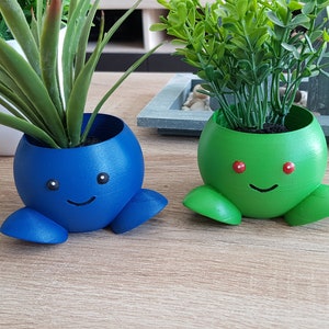 May include: Two 3D printed plant pots shaped like cartoon characters. The pot on the left is blue with a smiling face and black eyes. The pot on the right is green with a smiling face and red eyes.