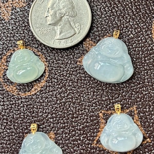 May include: Four translucent, light blue jade Buddha pendants with gold-colored bails. A U.S. quarter coin is included for size reference. The pendants are on a brown, textured surface.