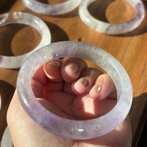 May include: A set of five amethyst crystal bracelets. The bracelets are a light purple color and have a smooth, polished finish. They are stacked on a wooden surface.