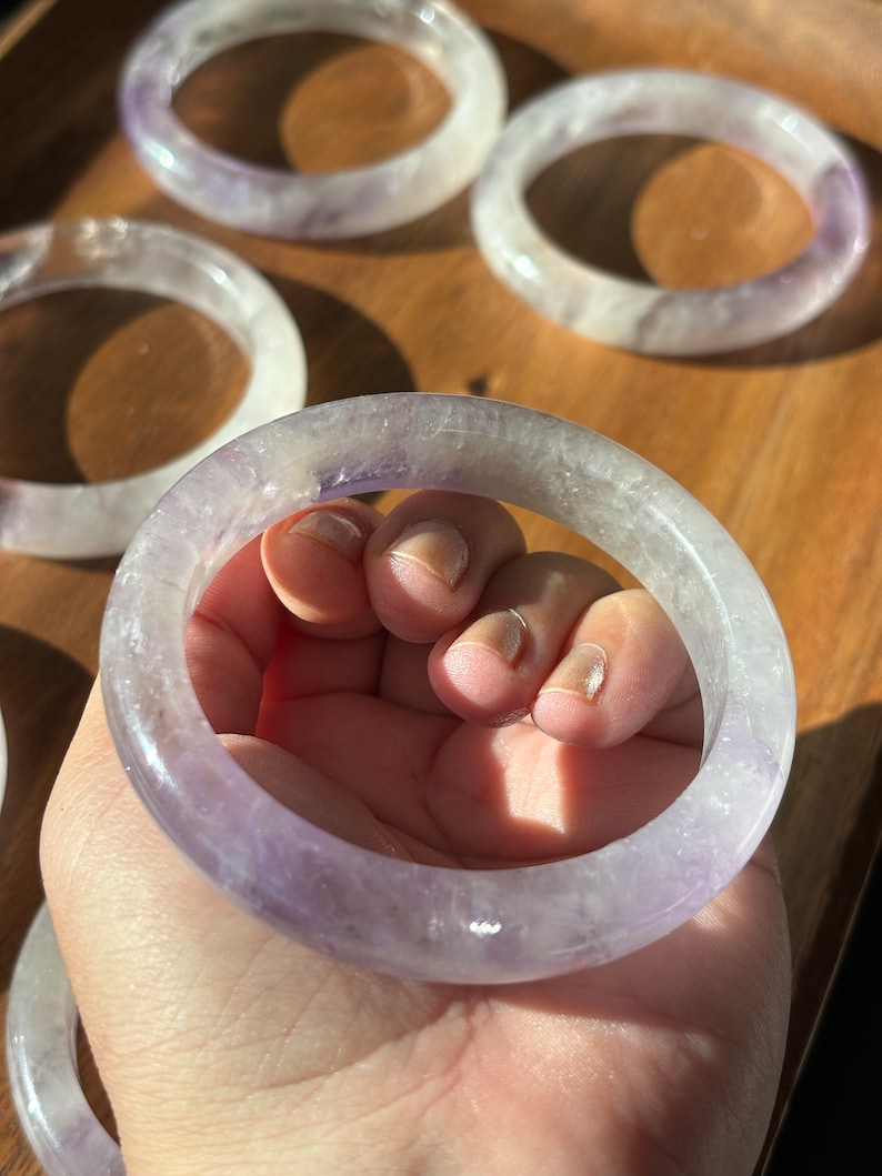 May include: A set of five amethyst crystal bracelets. The bracelets are a light purple color and have a smooth, polished finish. They are stacked on a wooden surface.