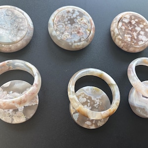 Puede incluir: Tres posavasos circulares de ágata flor y tres brazaletes a juego de ágata flor. Los posavasos son planos y redondos con patrones florales translúcidos en gris, blanco y rosa suave. Los tres brazaletes son lisos, pulidos y circulares.