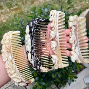 May include: Four decorative combs with intricate floral and nature-inspired carvings. One comb features a black and white marbled design. The other three combs are a light beige color, with detailed relief carvings of flowers and leaves.