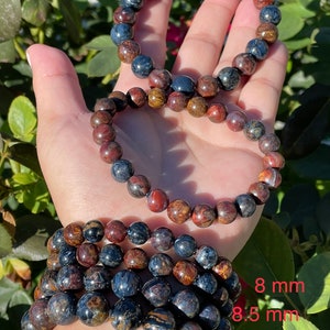 May include: Four bracelets made of tiger's eye beads in various sizes. The bracelets are stacked on a person's wrist. The beads are a mix of brown, gold, and blue colors. The sizes of the beads are labeled 8 mm, 8.5 mm, 10 mm, and 11 mm.
