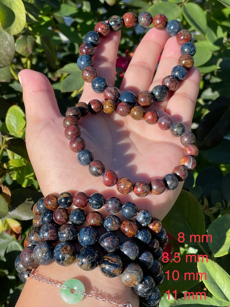 May include: Four bracelets made of tiger's eye beads in various sizes. The bracelets are stacked on a person's wrist. The beads are a mix of brown, gold, and blue colors. The sizes of the beads are labeled 8 mm, 8.5 mm, 10 mm, and 11 mm.