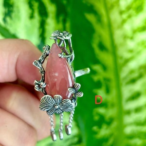 May include: A silver ring with a pink gemstone and floral details. The ring has a dangling chain with three charms.