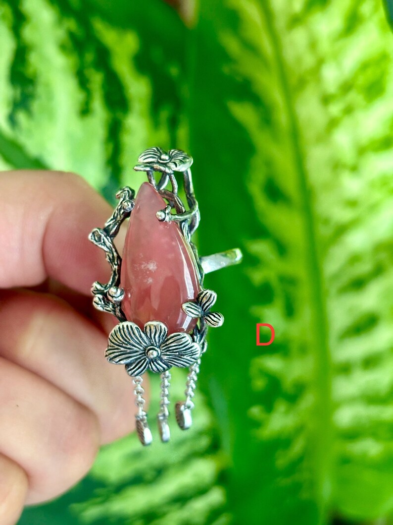 May include: A silver ring with a pink gemstone and floral details. The ring has a dangling chain with three charms.