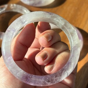 May include: A clear, translucent amethyst bangle bracelet with a light purple hue. The bracelet is made of natural amethyst and has a smooth, polished finish.
