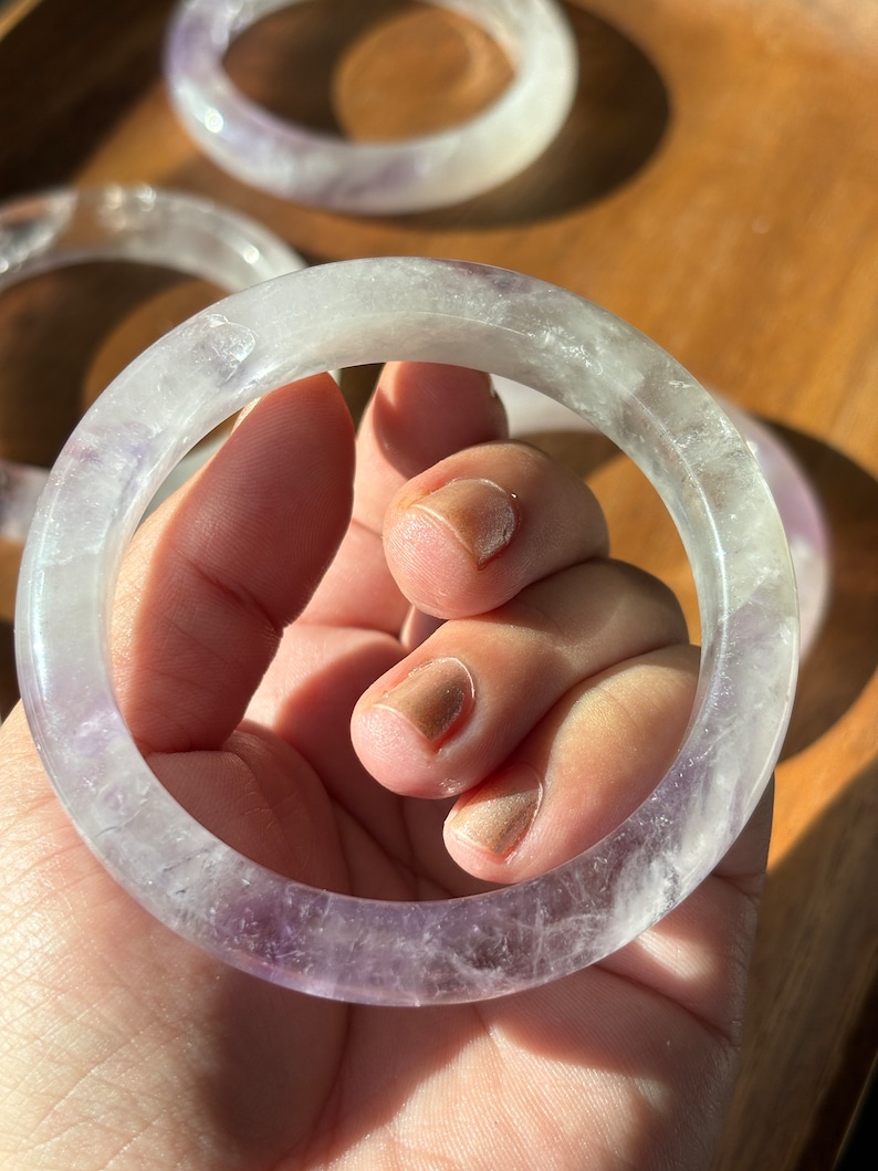 May include: A clear, translucent amethyst bangle bracelet with a light purple hue. The bracelet is made of natural amethyst and has a smooth, polished finish.