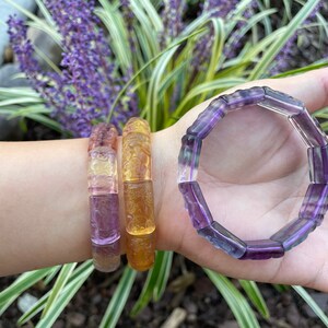 May include: Three bracelets made of carved gemstone beads. The bracelets are layered on a person's wrist. The top bracelet is purple, the middle bracelet is yellow and the bottom bracelet is purple and yellow.
