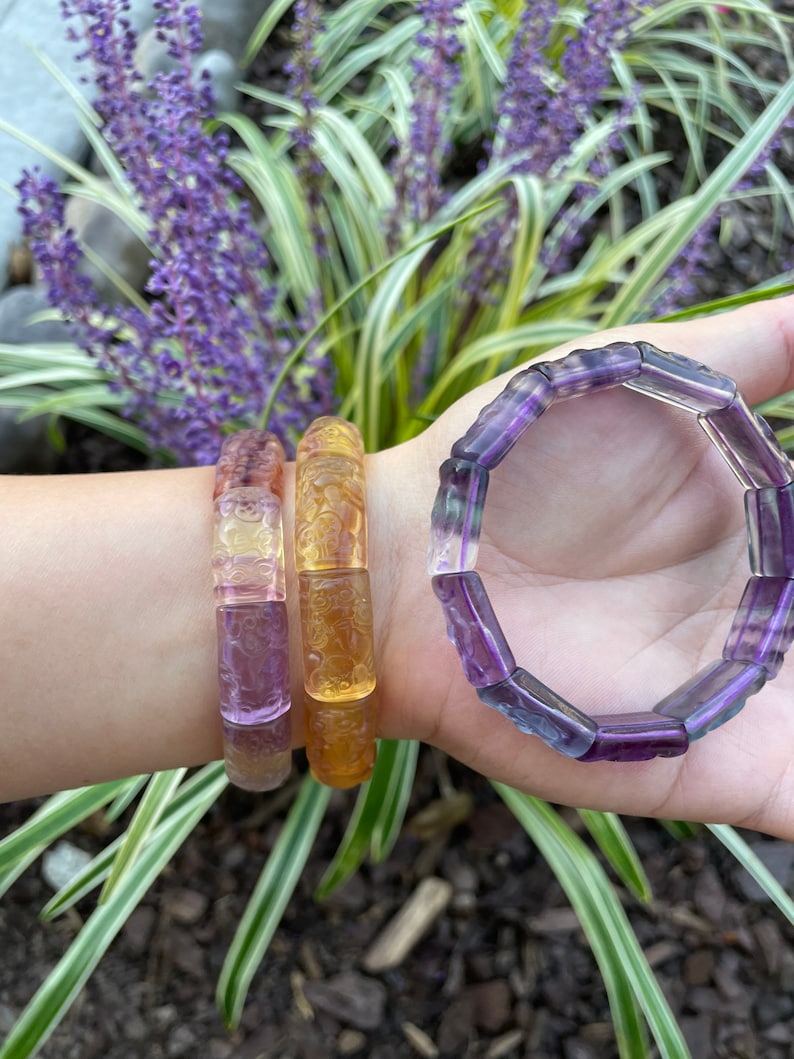 May include: Three bracelets made of carved gemstone beads. The bracelets are layered on a person's wrist. The top bracelet is purple, the middle bracelet is yellow and the bottom bracelet is purple and yellow.
