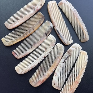 Puede incluir: Una colección de peines de cuerno de varios tamaños y formas. Los peines tienen un color marrón claro natural con vetas y patrones más oscuros. Los dientes están finamente espaciados y algunos peines tienen bordes decorativos tallados. Sobre una superficie oscura.