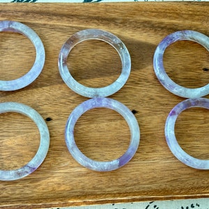 May include: Six amethyst gemstone bracelets, each with a light purple hue and a translucent appearance, are arranged in two rows of three on a wooden surface.