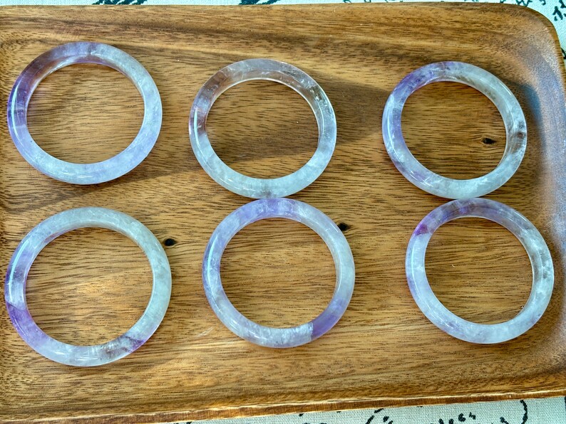 May include: Six amethyst gemstone bracelets, each with a light purple hue and a translucent appearance, are arranged in two rows of three on a wooden surface.