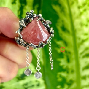 May include: A silver ring with a pink gemstone set in a floral design. The ring has three dangling chains with small charms at the end.