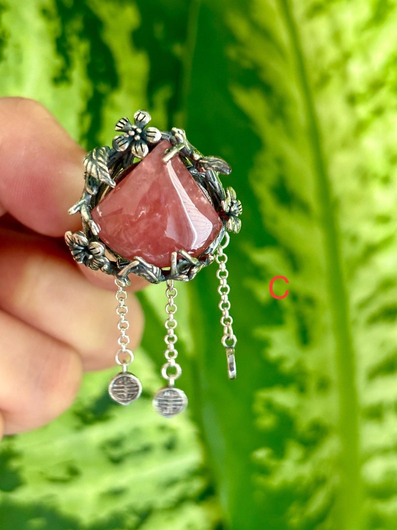 May include: A silver ring with a pink gemstone set in a floral design. The ring has three dangling chains with small charms at the end.