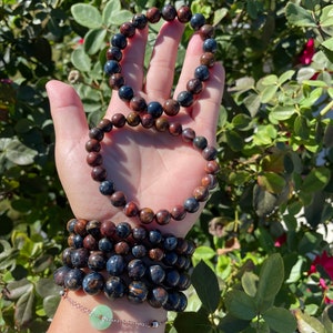May include: A stack of six bracelets made of dark blue and brown tiger's eye beads. The bracelets are layered on a person's wrist, with a silver chain bracelet with a green jade charm on top.
